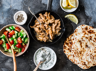 Ingredients for greek chicken gyros - fried chicken, tomato cucumber salad, tzatziki sauce and flatbread on a dark background, top view. Flat lay