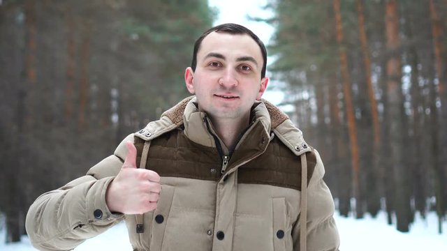 Portrait of man in a jacket is showing thumb up. Man stands in winter forest