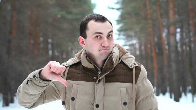 Portrait of man in a jacket is showing thumb down. Man stands in winter forest