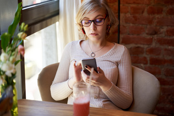 Serious adult woman with glasses sitting in a cafe, reading the news on the phone. A successful girl uses a smartphone in a restaurant, waiting for business partners to meet