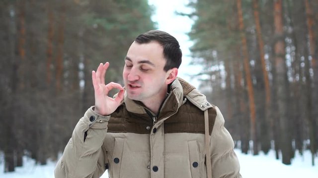 Portrait of handsome man in jacket is showing okay gesture at camera. Man stands in winter forest