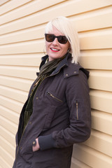 Medium vertical shot of pretty smiling platinum blonde young woman in dark olive coat and scarf wearing sunglasses seen leaning on exterior yellow wall with wood siding