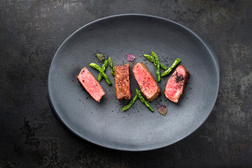 Barbecue dry aged wagyu fillet steak with blanched green asparagus tips and herbs as top view on a modern design plate with copy space