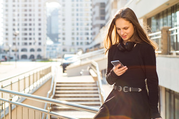 Outdoor portrait of young teenager brunette girl with long hair. girl on city in black dress looking on the smart phone in the street in a sunny day