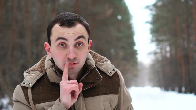 Portrait of young man in jacket is making silence gesture. Man stands in winter forest