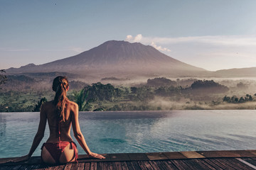 beautiful woman with dark hair in bikini posing near swimming pool with volcano Agung view