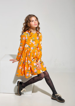 Fashion for girls 10 years in a yellow dress posing in the Studio on a cube. Curly hair, black shoes.