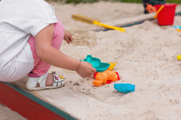 Little caucasian girl in a cap, t-shirt and striped leggings playing in the sandbox on the playground, close-up, day