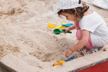 Fototapeta na wymiar Little caucasian girl in a cap, t-shirt and striped leggings playing in the sandbox on the playground, close-up, day