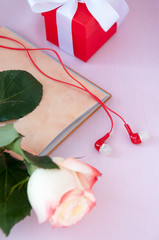 A light yellow-red rose lies on an open empty Notepad, headphones and a red box with a white ribbon on a pink background