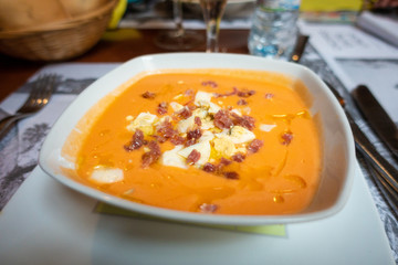 Closeup of a white ceramic bowl with salmorejo cordobes, a typical spanish tomato soup similar to the gazpacho, topped with serrano ham and croutons, on a set table