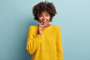 Enthusiastic pleased female model feels chilly, giggles positively, keeps fore finger on lips, being upbeat, satisfied to finish work, wears loose yellow jumper, isolated over blue wall. Happiness
