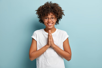 Friendly dark skinned female holds palms together over chest, shows namaste gesture, asks for help, has happy expression, wears white t shirt, optical glasses, isolated over blue background. - obrazy, fototapety, plakaty