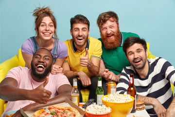 Group of diverse friends cheer as favourite team wins, show thumb up gesture, eat tasty pizza and popcorn, smile broadly, drink beer, isolated over blue background. People, entertainment, fun concept