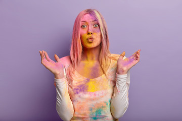 Happy Holi Day. Pleasant looking woman has folded lips, spreads hands, wears casual clothes, has glad expression, dirty with colorful powder, isolated over lilac studio wall. Colored dust explode