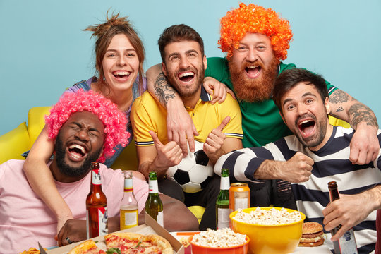 Shot of friendly companions embrace and smile happily, cheer with winning favourite team, have nice time together watching exciting football game, drink beer and eat fast food. Funny fans support