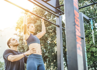 Obraz premium Personal trainer helping his friend to do pull up during the calisthenics session - Couple doing sport together in the park at sunset