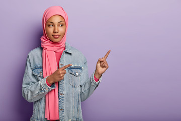 Photo of self assured Arabian lady in traditional pink hijab, shows you right way, points at blank space with both fore fingers, invites going there, promots copy space. Check it out with me