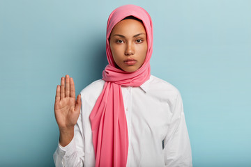 Slow down and stop. Serious dark skinned Arabic woman shows refusal gesture, raises palm towards camera, expresses refusal, wears pink headwear and white shirt, isolated over blue background.