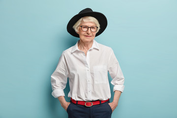 Fashionable mature woman wears stylish black hat, white shirt and trousers with red belt, keeps...