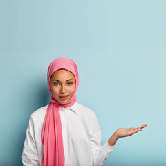 Religious confident Muslim woman seller raises palm over blank space, holds invisible object, wears...