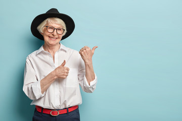 Image of attractive wrinkled woman with appealing look, feels refreshed, young for her age, points at upper right corner, satisfied with product, wears white shirt, black headgear, optical glasses