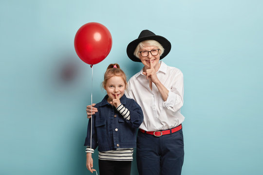 Photo of smiling small cute girl and senior woman demonstrate hush sign, tell secret, have glad expression, feel excited, come on festive event, isolated over blue background. Shush, be quiet