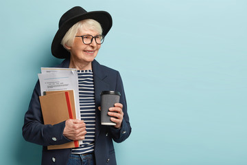 Indoor shot of elderly lady in high spirit, finishes paper work has coffee break, wears hat and...