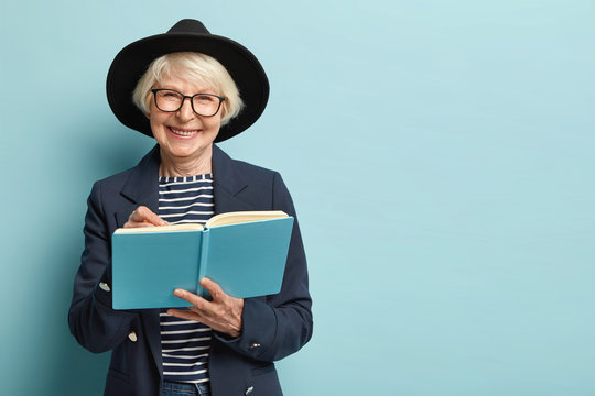 Portrait of pleased female pensioner writes plan strategy in diary, has nice clever look, wears glasses and black hat, isolated over blue studio wall with empty space. Businesswoman with notepad