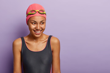 Isolated shot of dark skinned woman athelete covers distance swimming, has healthy dark wet skin, wears swimsuit, goggles and hat, looks gladfully aside, trains in swimming pool. Recreation time