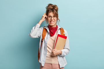 Obraz premium Puzzled schoolgirl holds notebook and spiral diary, bites lips, scratches head as recollects information in mind, wears stylish bandana, transparent glasses, rucksack on back. Studying concept