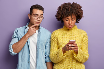 Diverse woman and man stand indoor, dark skinned lady focused in smart phone, types feedback, sends text message, curious guy peeks in screen, wears denim shirt. Multiracial relationship concept