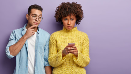 Curious man peeps in cell phone of girlfriend. Angy woman with Afro haircut, checks notification on gadget, wears yellow jumper, stand next to each other over purple background. Mixed race partners