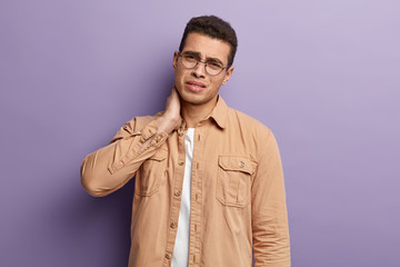 Health problem concept. Frustrated young Caucasian man has neck pain, looks with sad expression, wears brown shirt, round spectacles, needs massage, tilts head, isolated over purple background.