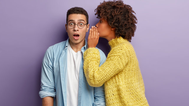 Photo of Afro American woman whispers secret to boyfriend, tells shocking news. Astonished young man keeps jaw dropped from surprisement, hears terrible rumors, isolated over purple background