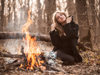 Attractive young woman with black cape sitting in forest near bonfire.