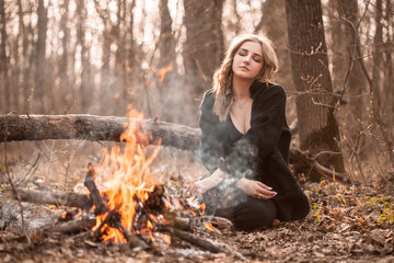 Attractive young woman with black cape sitting in forest near bonfire. Eyes closed