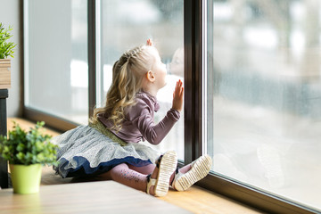 A beautiful child is sitting near a large window. A little girl, 4-5 years old, is sitting on the windowsill of a large window in  cafe.