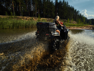 Beautiful blonde girl on a black ATV rides on the river, standing on the mountain. Around the forest, sand and blue sky.She rides on a green swamp, in the mud and all wet. She's wearing a blue bikini. © Pavel
