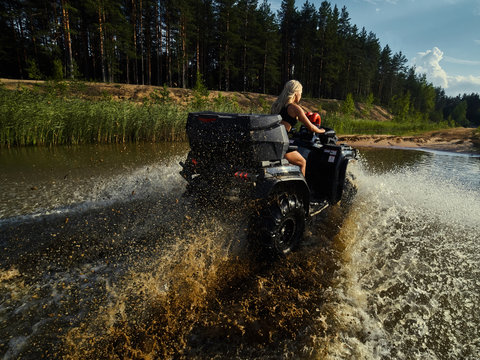 Beautiful blonde girl on a black ATV rides on the river, standing on the mountain. Around the forest, sand and blue sky.She rides on a green swamp, in the mud and all wet. She's wearing a blue bikini.