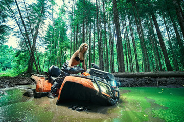 Piękna blondynki dziewczyna na orang ATV jedzie na rzece, stoi na górze. Wokół lasu, piasku i niebieskiego nieba, jeździ na zielonym bagnie, w błocie i cała mokra. Ona ma na sobie niebieskie bikini. - 264766290