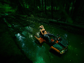Piękna blondynki dziewczyna na orang ATV jedzie na rzece, stoi na górze. Wokół lasu, piasku i niebieskiego nieba, jeździ na zielonym bagnie, w błocie i cała mokra. Ona ma na sobie niebieskie bikini. - 264766424