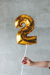 Gold number two balloon in a female hand on gray wall background isolated