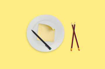 Blank square paper sticker with curved corner in white round plate near wooden pencil and black plastic pen in center of yellow table. Top view. Menu template, business or education concept