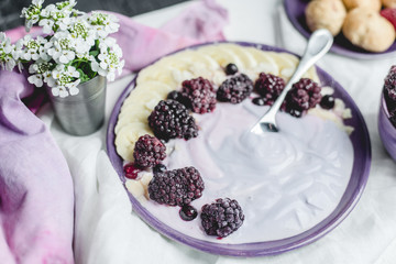 fruity yogurt with banana slices, frozen forest fruits and almond nut flakes on a plate surrounded by frozen berries and sweet buns