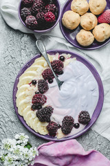 fruity yogurt with banana slices, frozen forest fruits and almond nut flakes on a plate surrounded by frozen berries and sweet buns