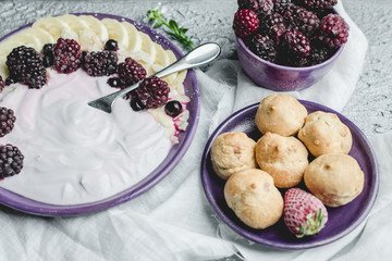 fruity yogurt with banana slices, frozen forest fruits and almond nut flakes on a plate surrounded by frozen berries and sweet buns