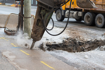 A large pneumatic jackhammer breaking a frozen road and sidewalk. Bits of concrete fly from the...
