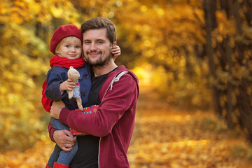 Fototapeta premium Father hugs and kisses his daughter in autumn day. The girl is holding a teddy bear. They look at the viewer.