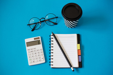 Top view of blue office desk, with cup of coffee, empty notebook and pencil, glasses and white calculator.